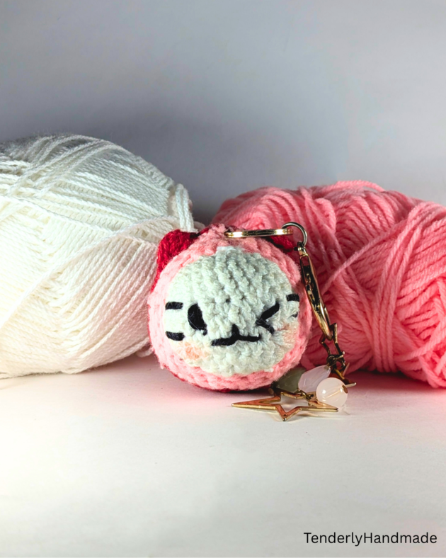 A round pink and white crochet Meowchi cat charm with a gold star keychain, placed in front of white and pink yarn balls.