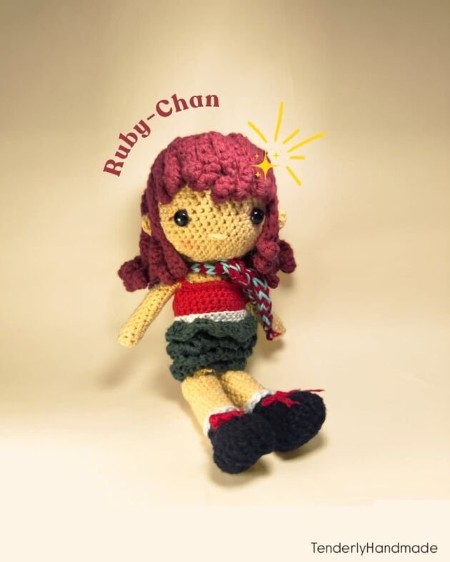 A handmade crochet doll named Ruby-Chan with curly red hair, a red top, a dark green skirt, and a patterned scarf sitting on a neutral background.