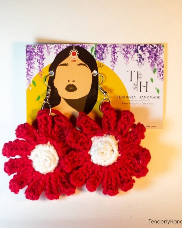 A pair of large red and white crochet dahlia flower earrings displayed on a Tenderly Handmade branding card featuring a woman in traditional attire.