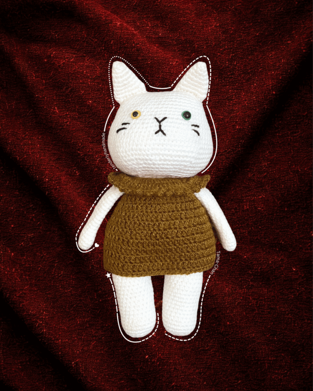 Frontal view of the Tama-Chan cat doll with mismatched eyes and black whiskers lying on a dark red textured background.
