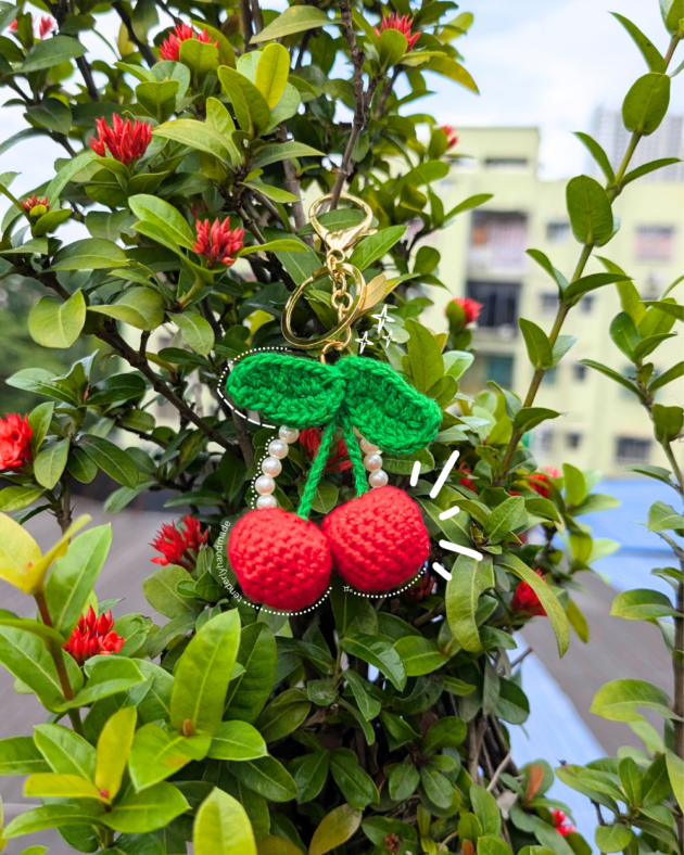 A bright red crochet cherry charm with pearl strands hanging from a green garden plant, highlighting its vibrant colors.