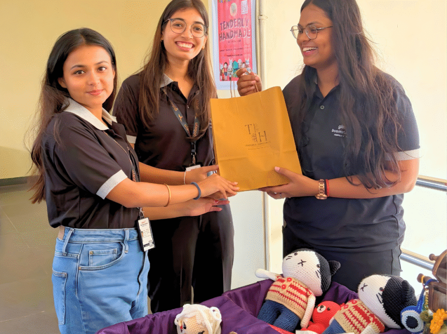 Two women in matching black Tenderly Handmade polo shirts smiling while holding a brown branded paper shopping bag and a small crochet bear keychain.