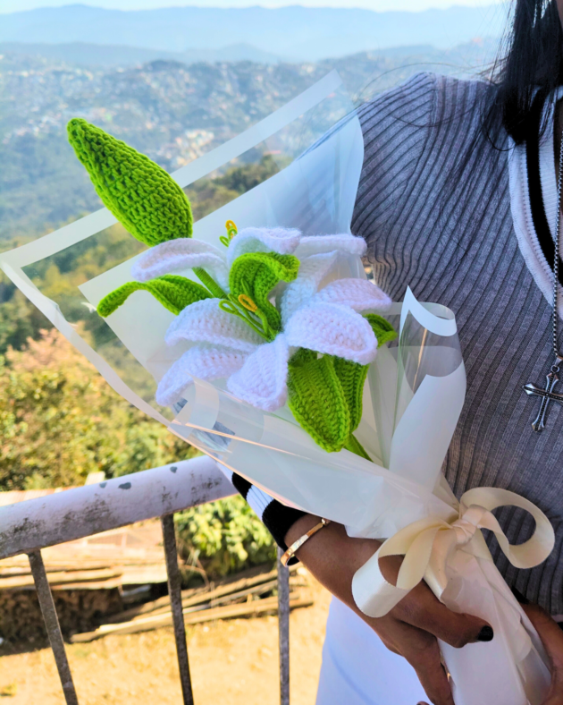 Close-up of a realistic white crochet lily bouquet with green leaves and a yellow stamen, wrapped in elegant white paper.