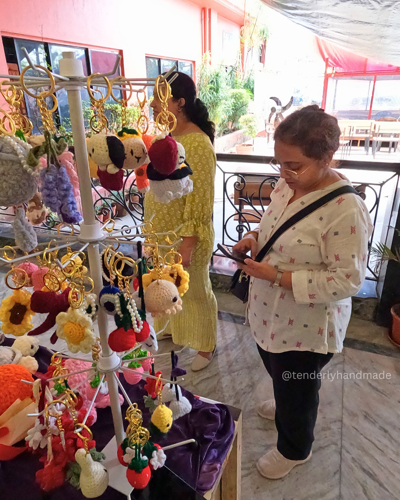 Customers examining colorful handmade crochet plushies, including octopuses and a green dinosaur, at a table display inside a college corridor.