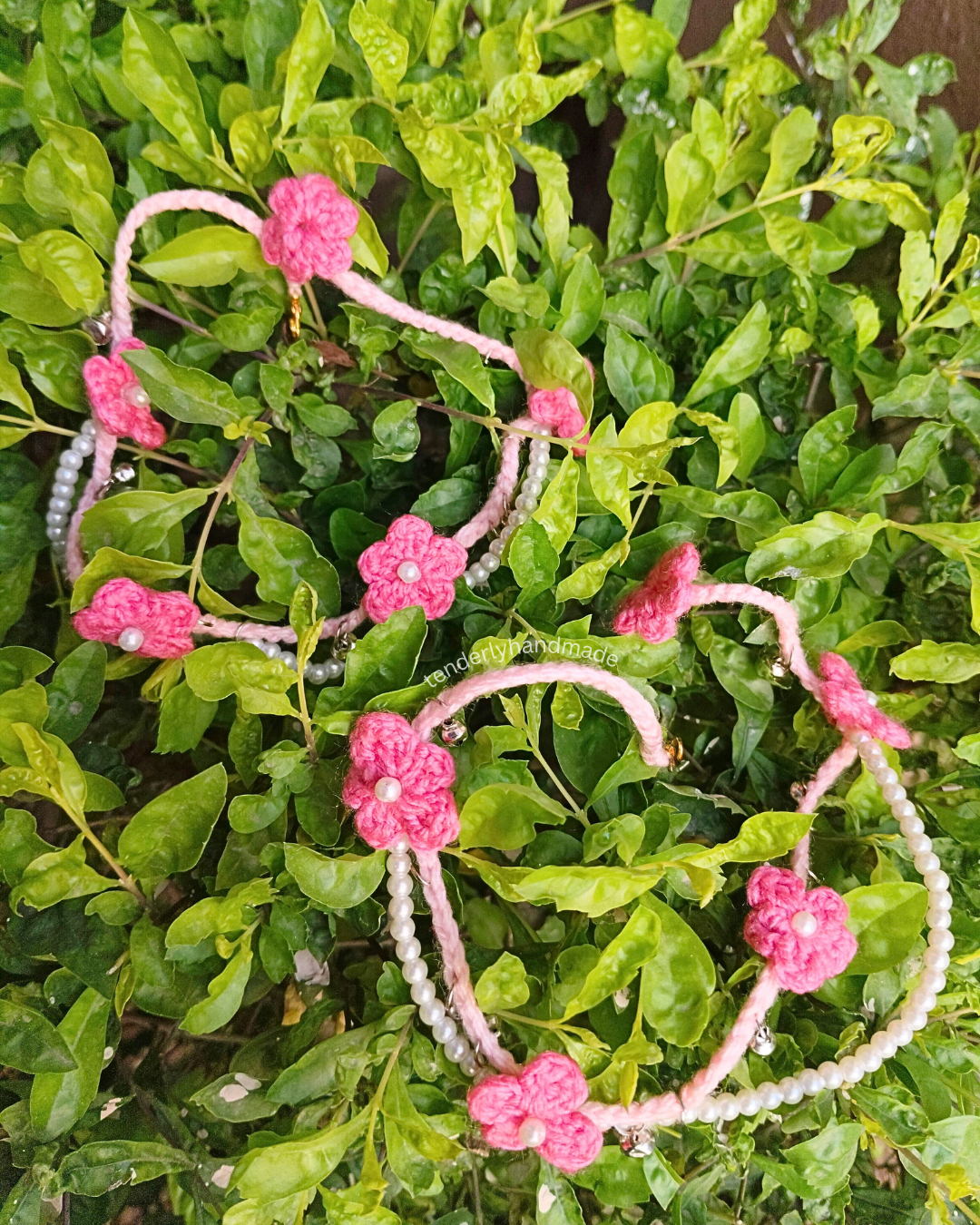 Overhead view of two pink crochet payals adorned with tiny flowers, white pearl beads, and silver ghungroos spread on lush green leaves.